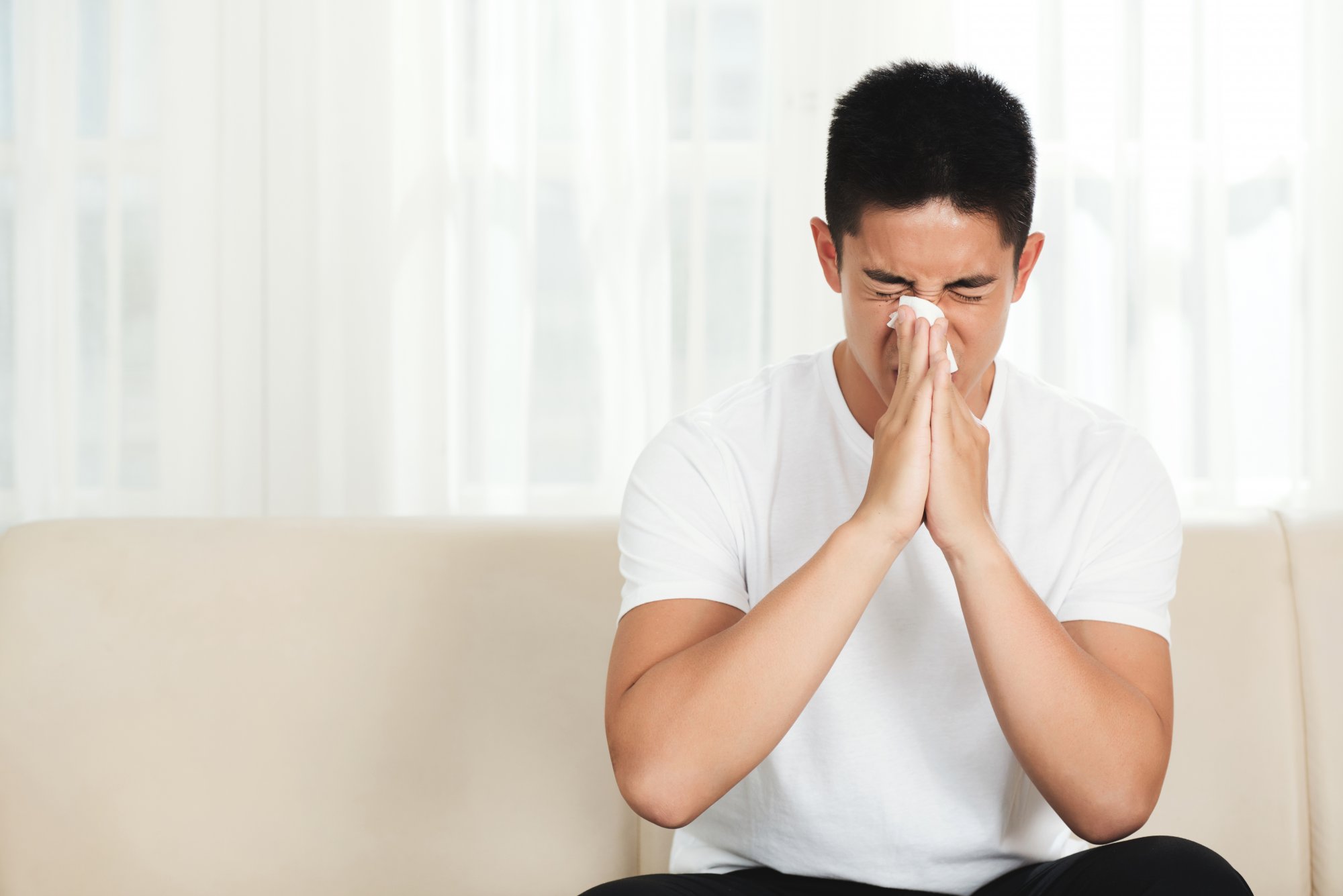 young-asian-man-sitting-couch-home-blowing-nose-with-tissue